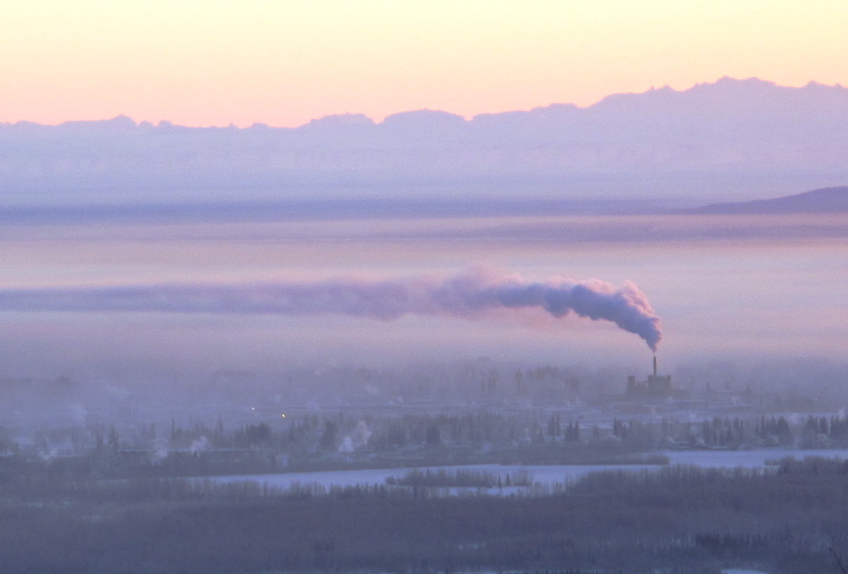 Sunshine lights fog in a wide valley with mountains in the background. A plume from a power plant rises above the fog.